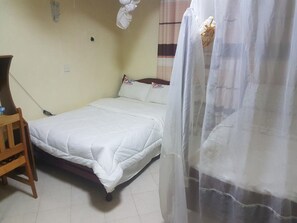 Habitación individual con una cama, una silla de madera, una mesita de noche y una cortina.