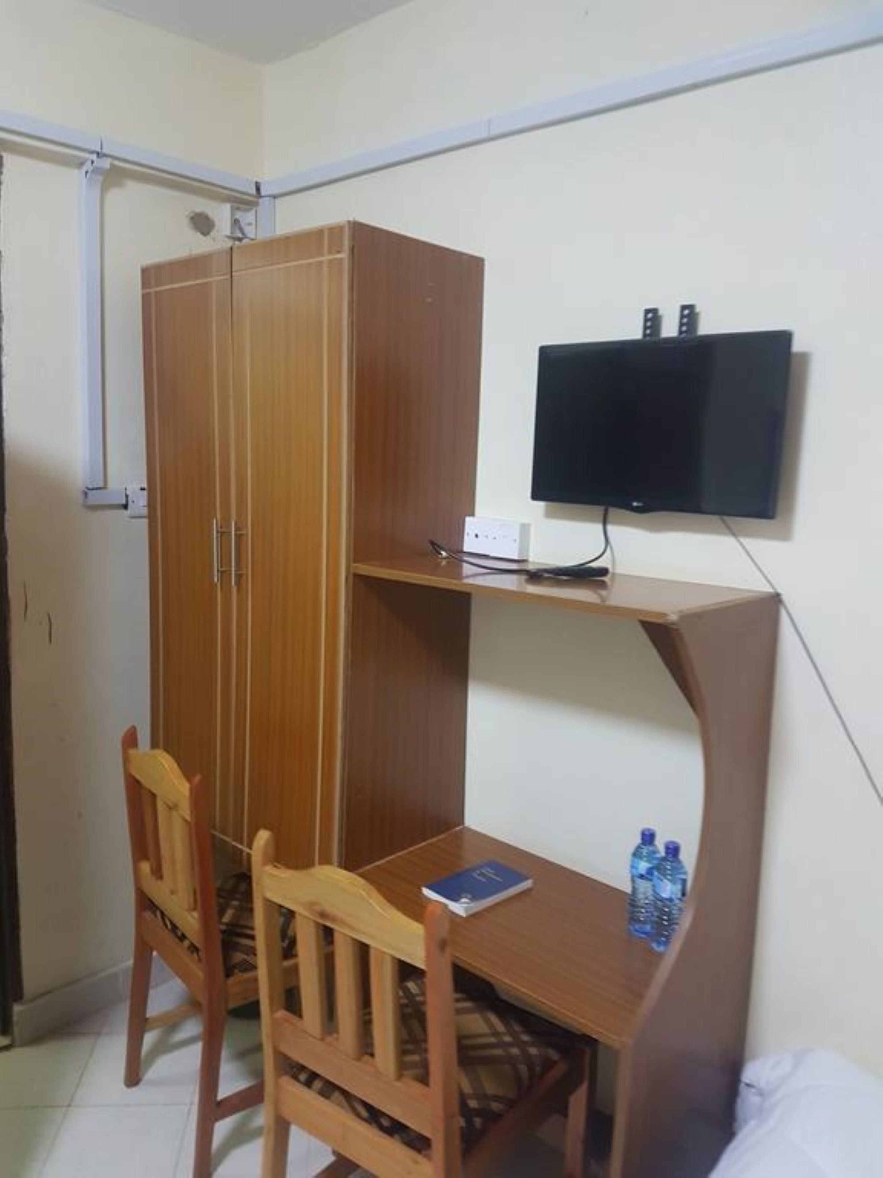 Ein Zimmer mit einem Holzkleiderschrank, einem Schreibtisch mit Stuhl, einem Wand-Flachbildfernseher und einer Wasserflasche auf dem Schreibtisch.