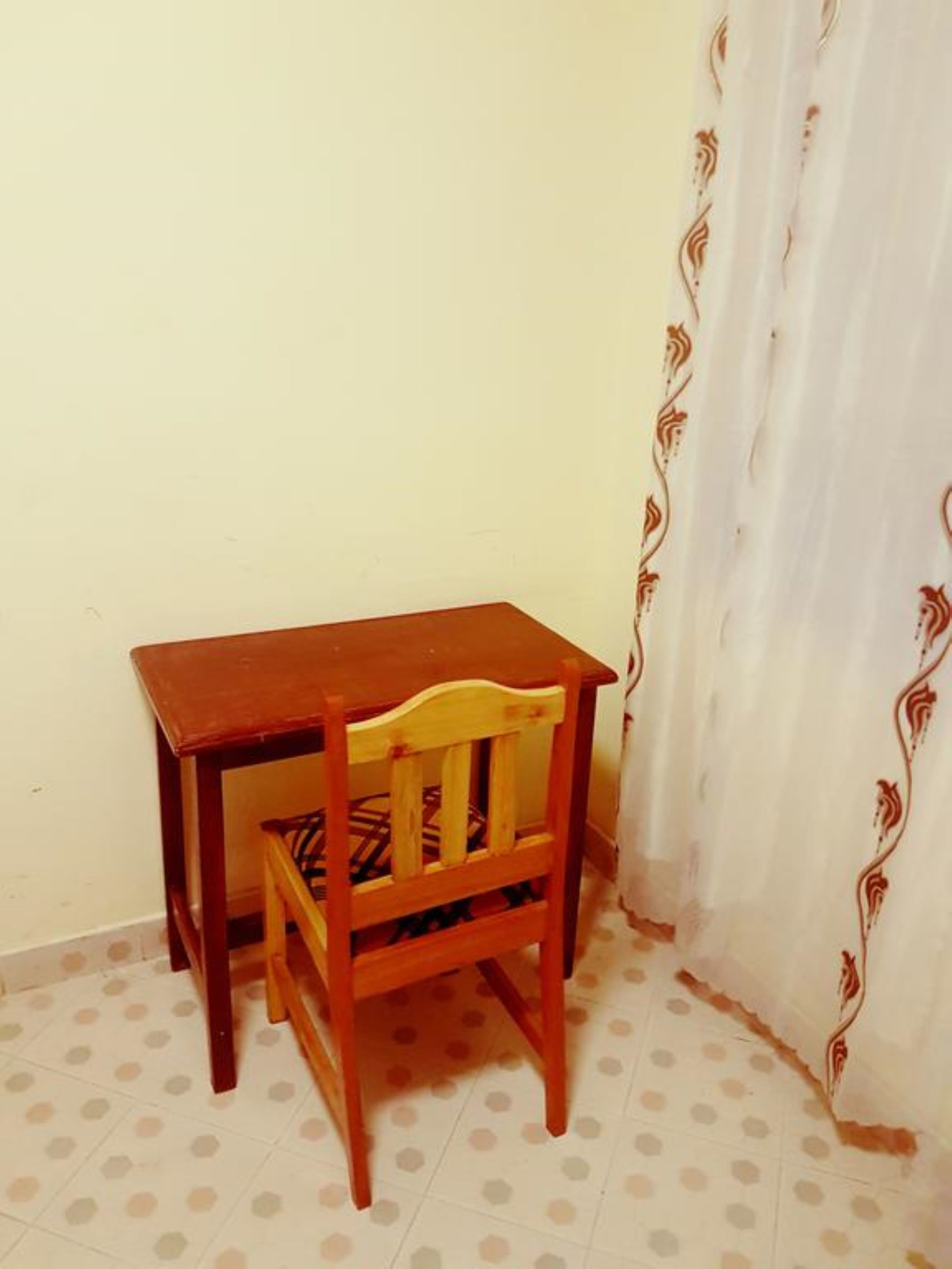 A small wooden table with a chair in a corner room with a polka dot floor and a white curtain with a brown pattern.