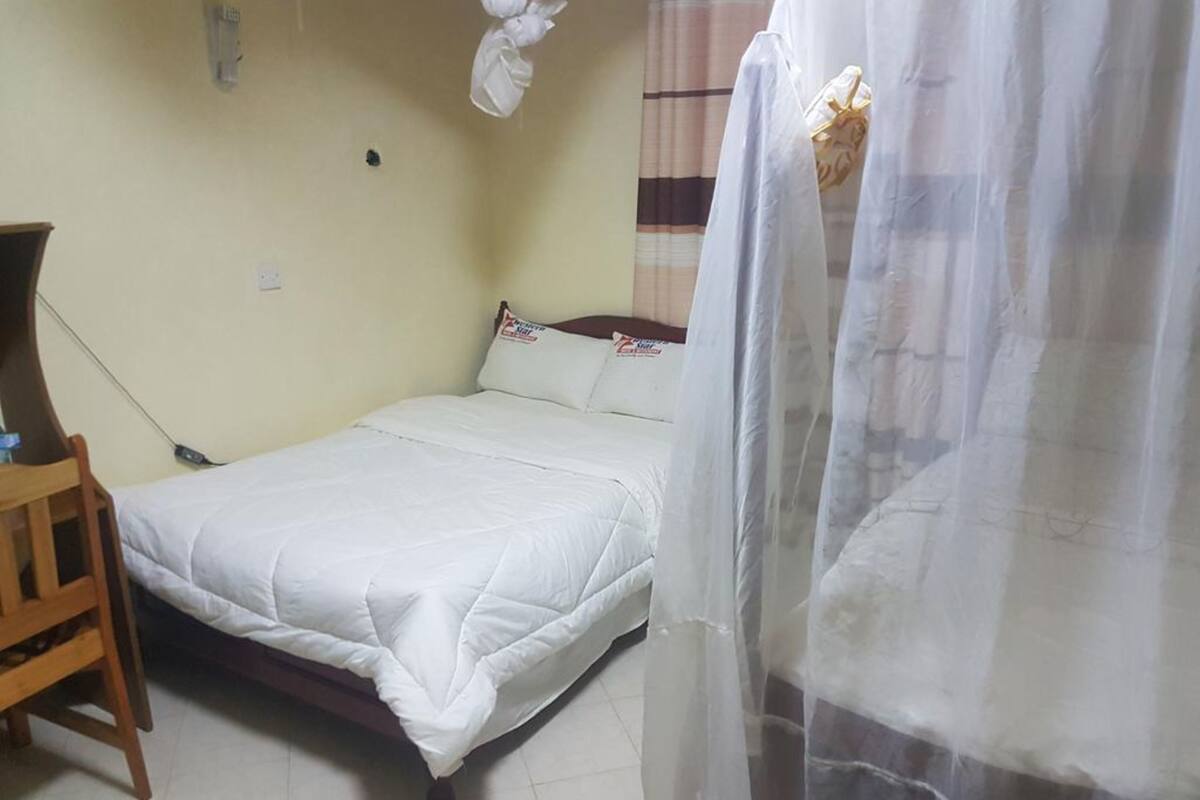 A single bed with a white bedspread, a wooden chair, a bedside table, and a curtain.
