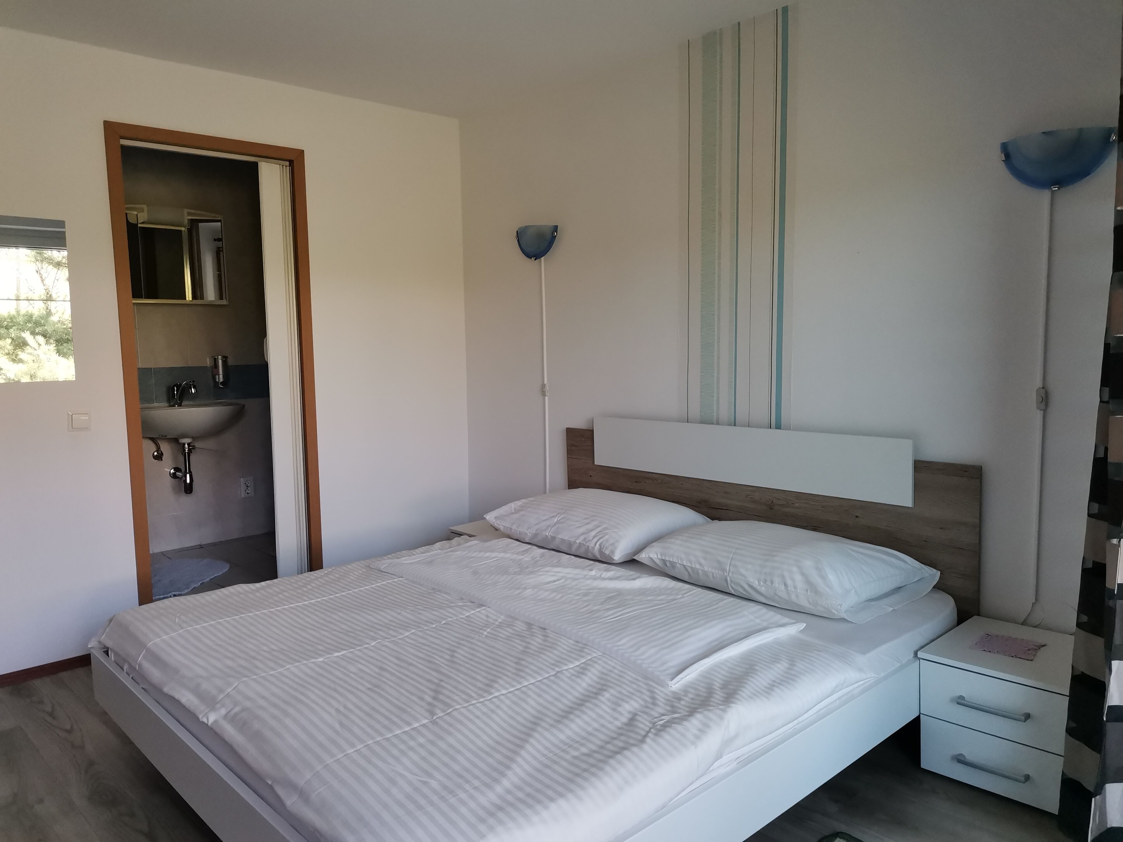 Un dormitorio con cama, mesita de noche y velador. Se ve una ventana con vistas a árboles.