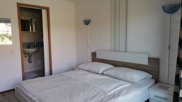 Un dormitorio con cama, mesita de noche y velador. Se ve una ventana con vistas a árboles.