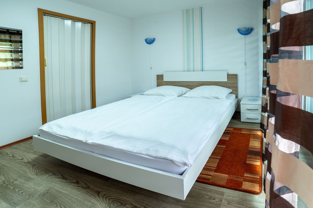 A modern bedroom with a single bed, a nightstand, a wall-mounted lamp, and a window with blinds.