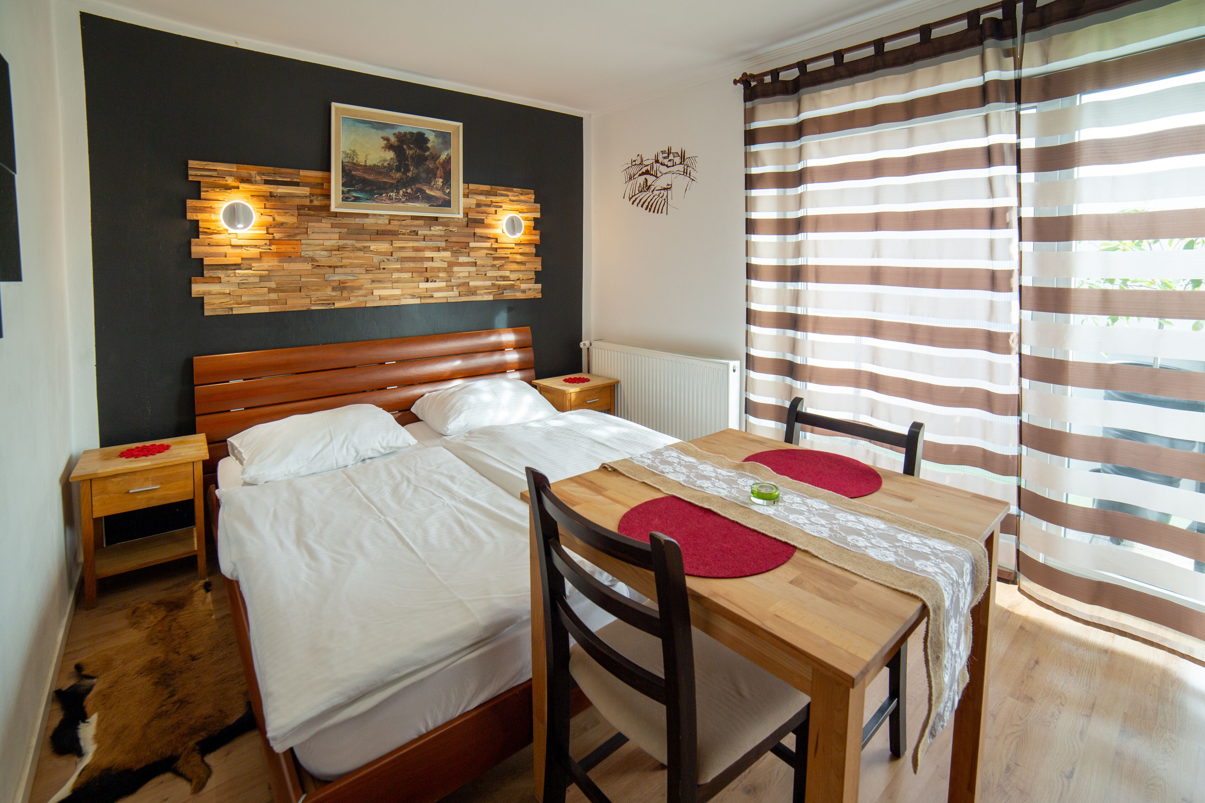 A bedroom with a wooden bed, a bedside table, a wooden dining table with chairs, a red rug, and a cityscape wall art.