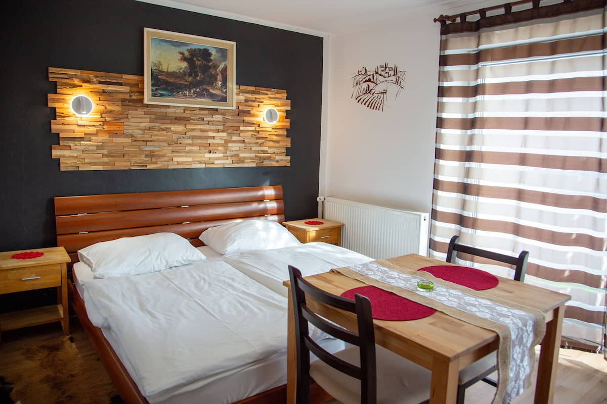 A bedroom with a wooden headboard, a bed with white linens, a wooden nightstand with a red flower, a wooden table with chairs, and a window with striped curtains.