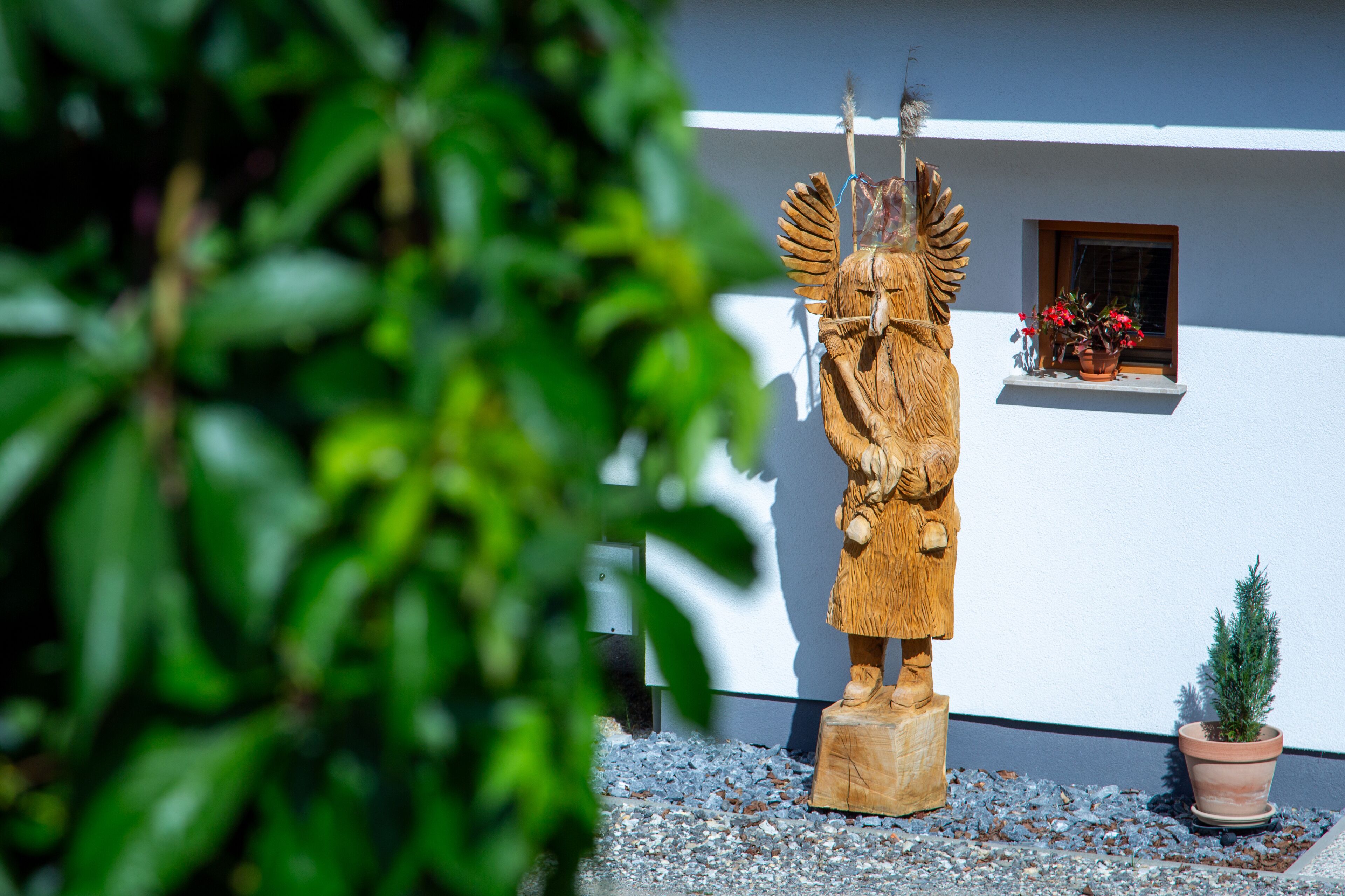 Una escultura de madera de una figura con alas, un báculo y un pájaro posado en su hombro, ubicada sobre un pedestal frente a un edificio blanco con una ventana y una planta en maceta.
