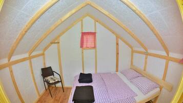 A small, attic-style room with a sloped ceiling, a single bed with a patterned bedspread, a chair with a cushion, and a small window with a checkered curtain.