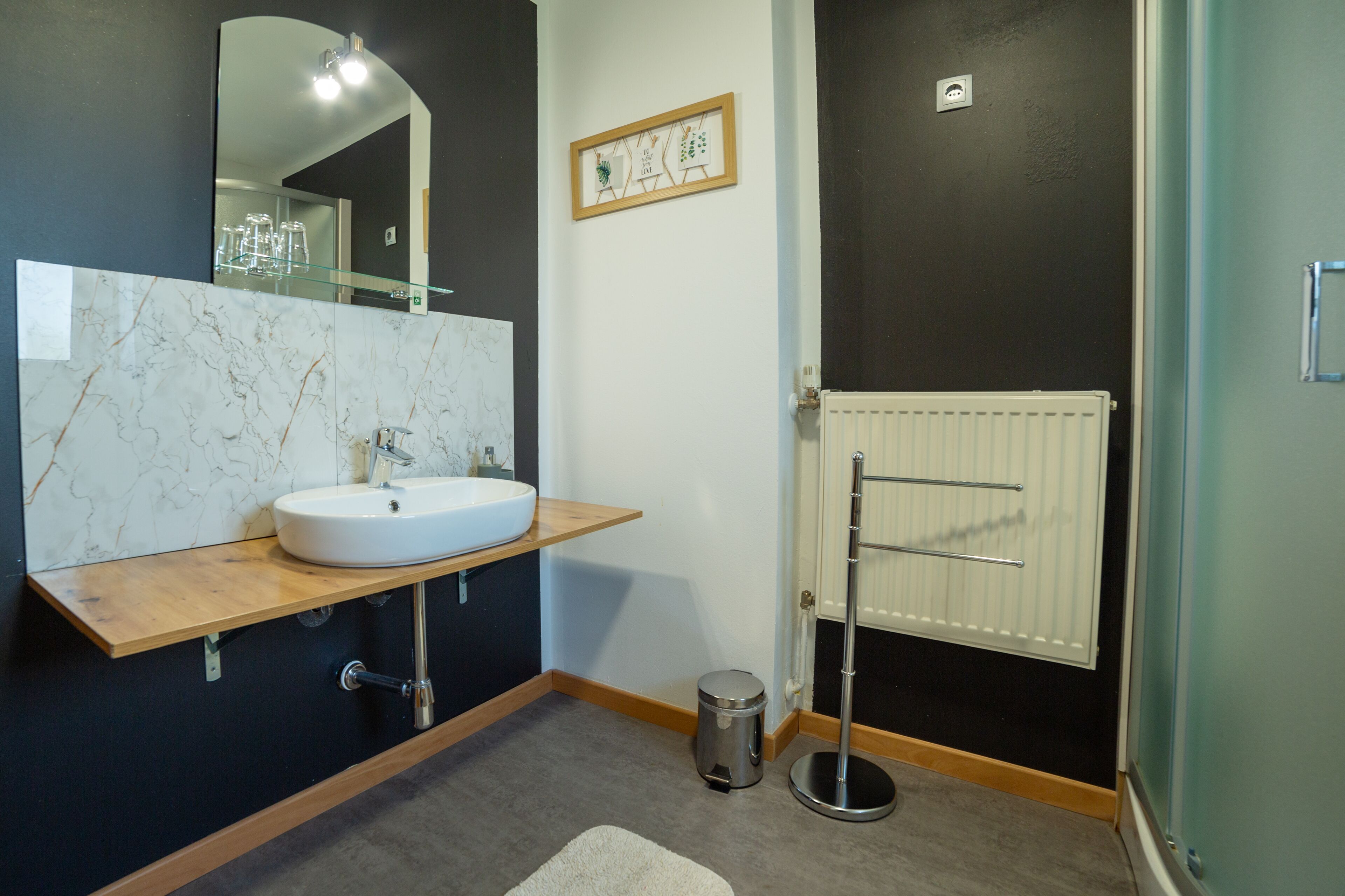 A modern bathroom with a marble sink, a wooden vanity, a mirror, a radiator, and a small trash bin.