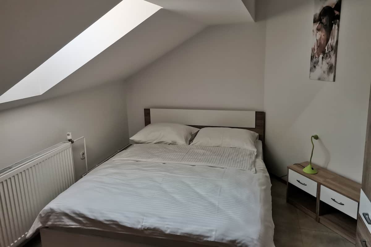 A bedroom with a sloped ceiling, a bed with white bedding, a radiator, a nightstand with a lamp, and a wall-mounted artwork.