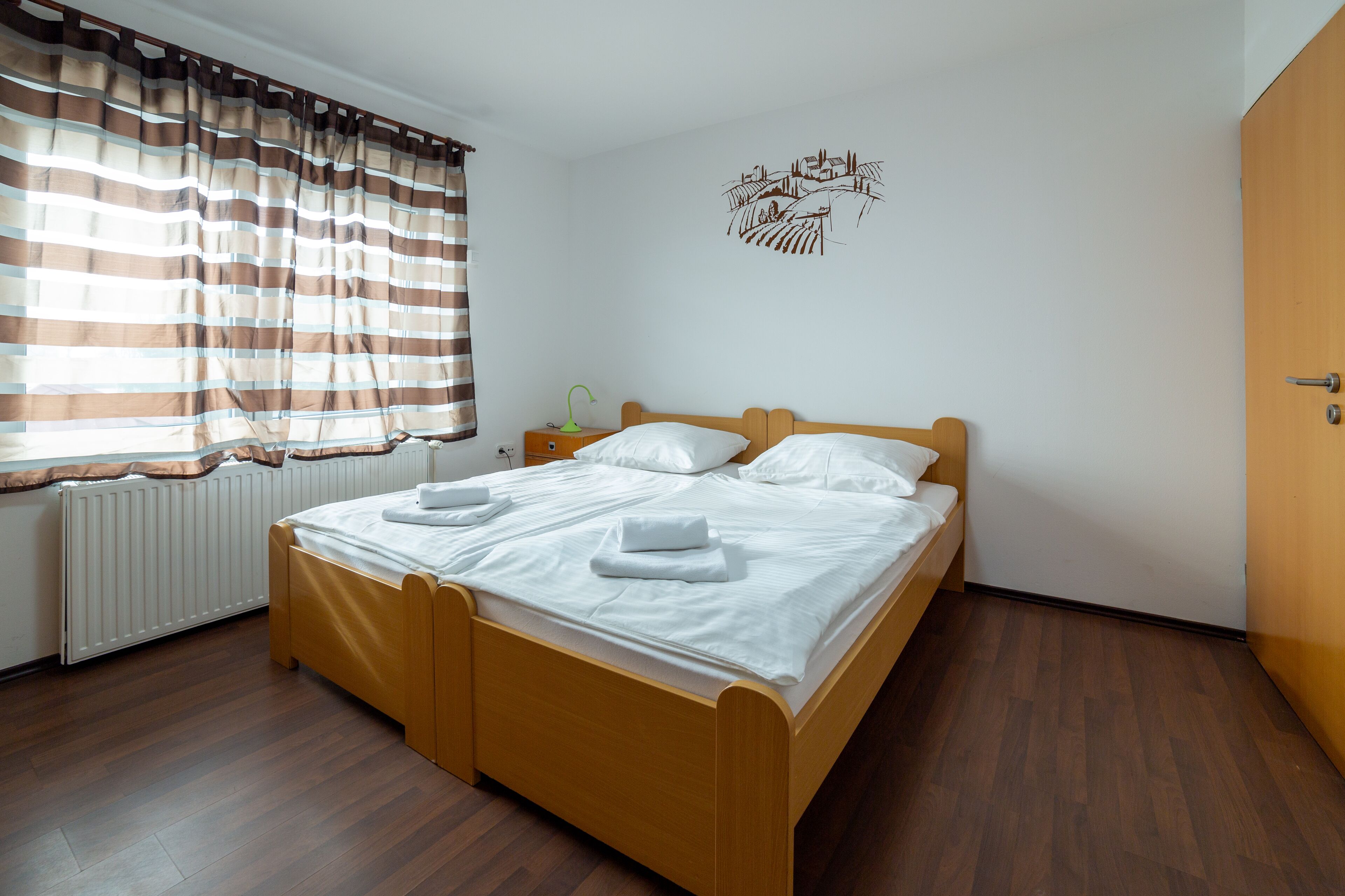 A neatly made double bed with white linens and towels, a bedside table with a lamp, and a window with striped curtains.