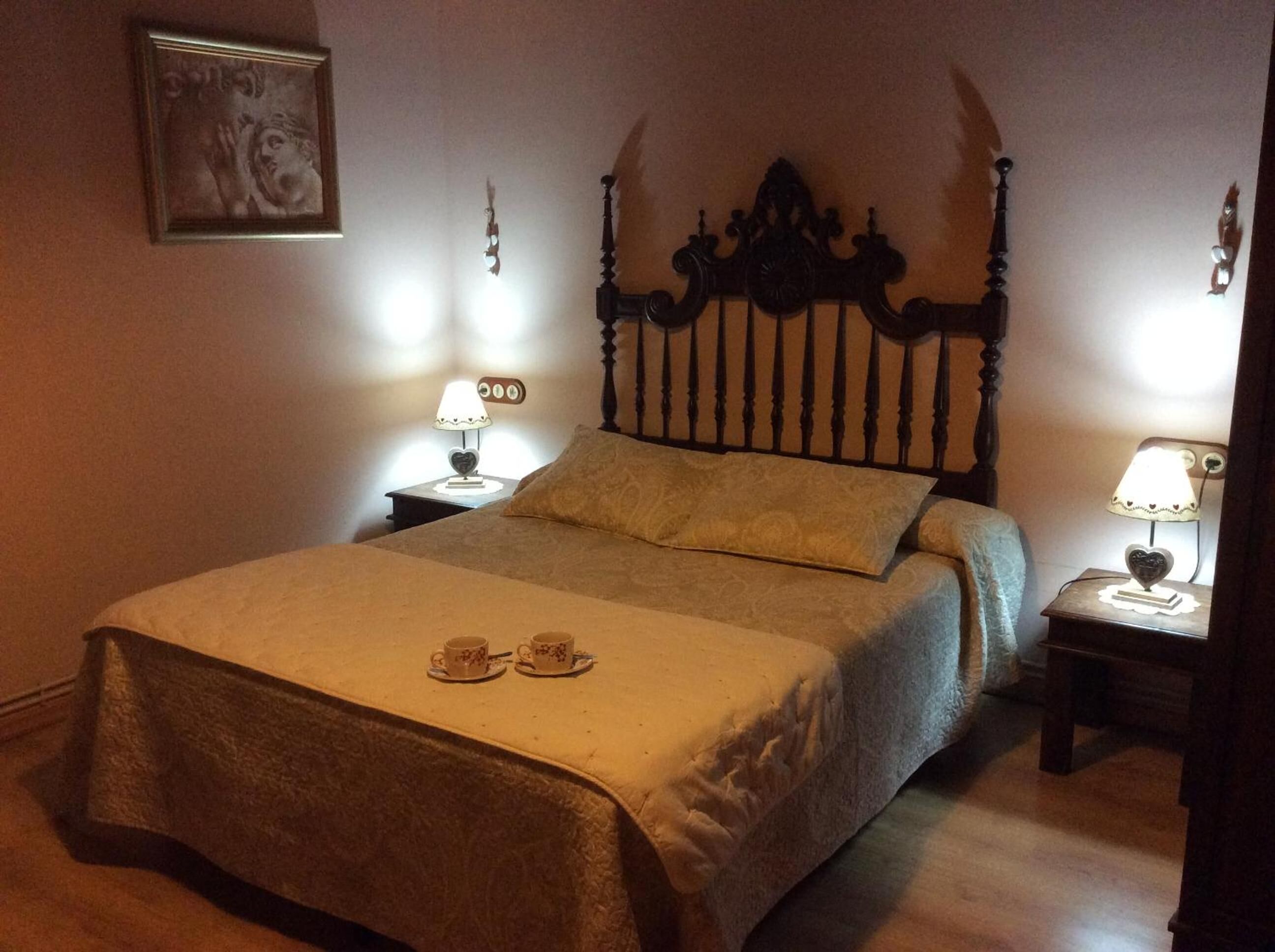 Un lit bien fait, recouvert d’une couvre-lit beige, et sur lequel reposent deux tasses décoratives. Deux tables de chevet, chacune équipée d’une lampe, sont placées de part et d’autre du lit.