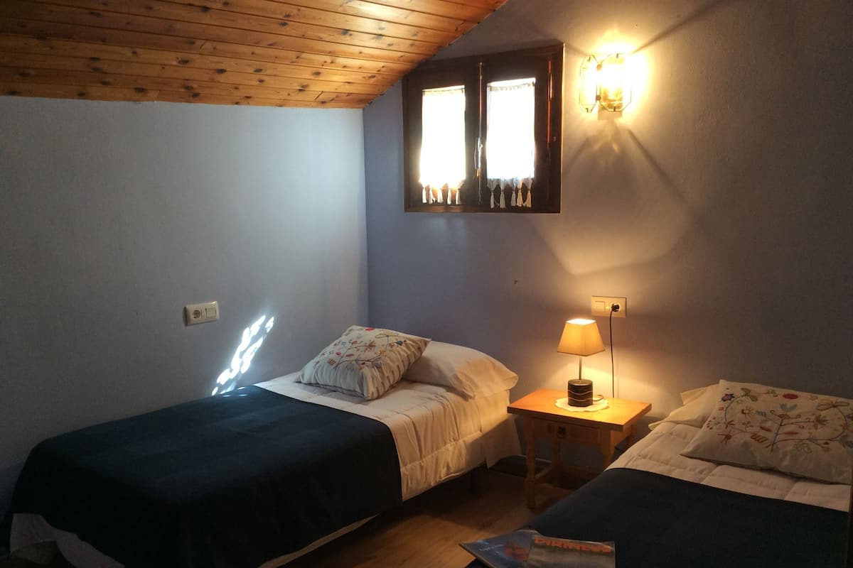 A room with two beds, a nightstand with a lamp, and a window with curtains.