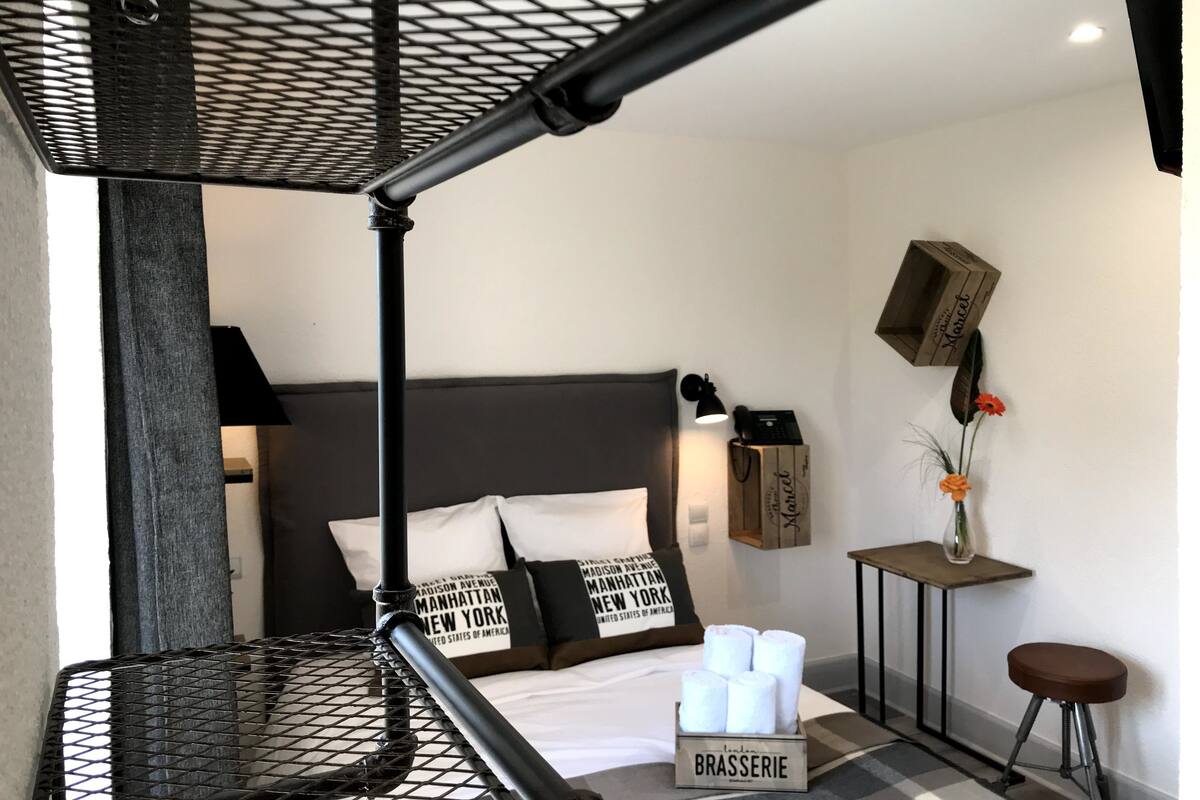 Moderni hotellihuone, jossa on sänky, pieni pöytä lampun kanssa, tuoli ja laatikko, jonka päällä on teksti ”BRASSERIE”.
