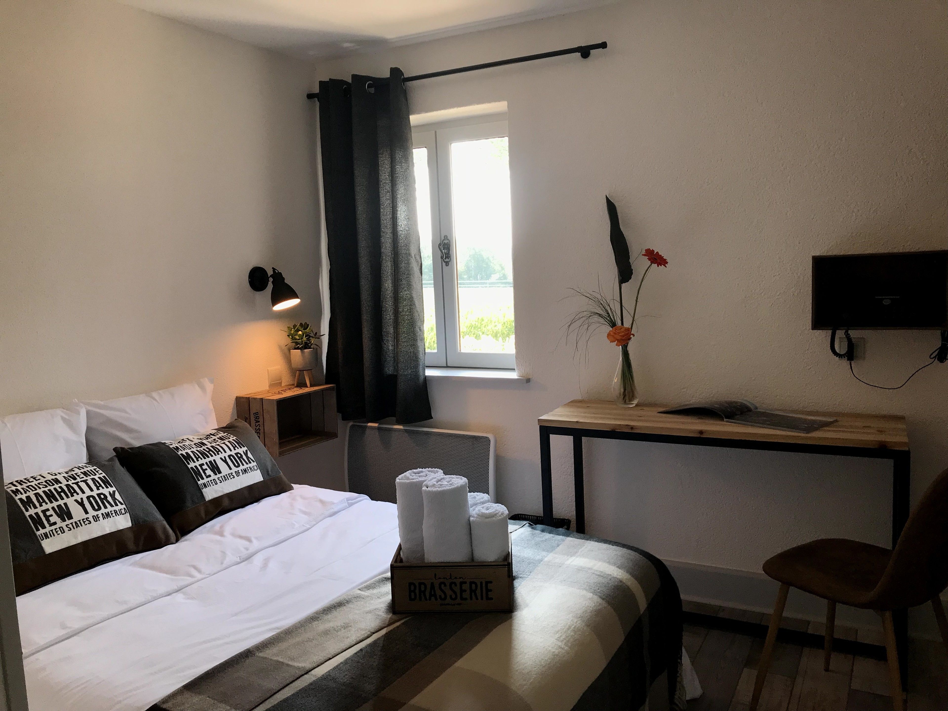 Habitación de hotel con una cama, una mesita con un florero, una silla, un escritorio y un televisor.
