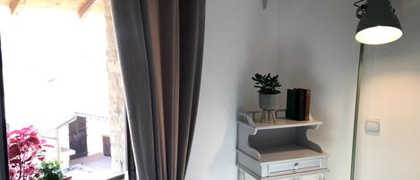 Una habitación con ventana, cortinas, un estante blanco pequeño con una planta y libros, una lámpara de pie y una pared blanca.