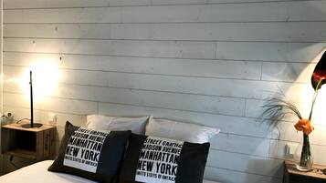 A hotel room with a bed, two pillows with 'Manhattan New York' text, a nightstand with a lamp, and a vase with a flower.