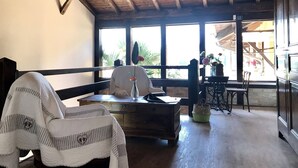 Una habitación acogedora con techo de madera, ventanas amplias, una silla blanca, una mesa de madera y una cesta verde.