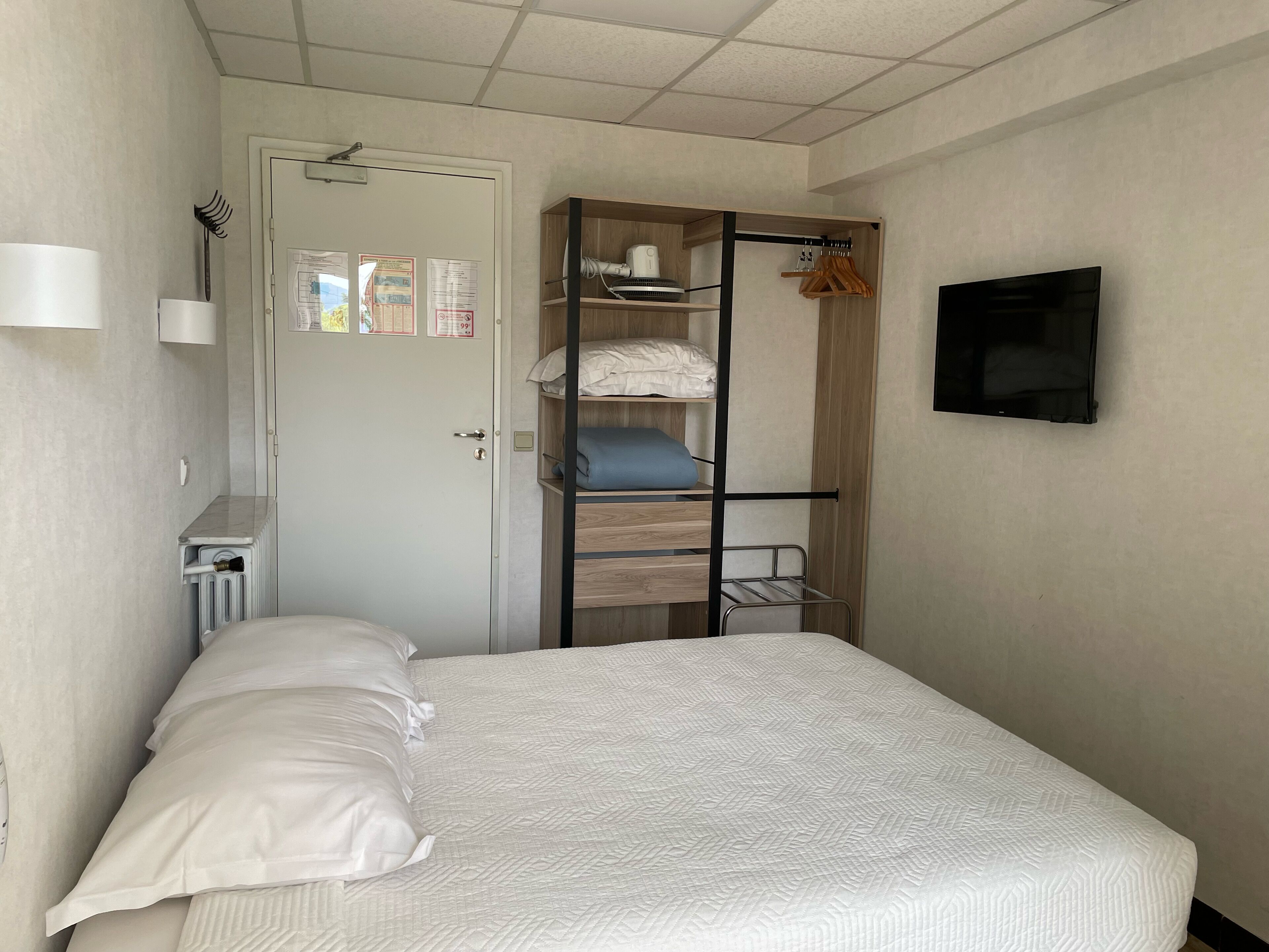 A bedroom with a bed, a wooden bunk bed, a radiator, a TV, and a door with notices.