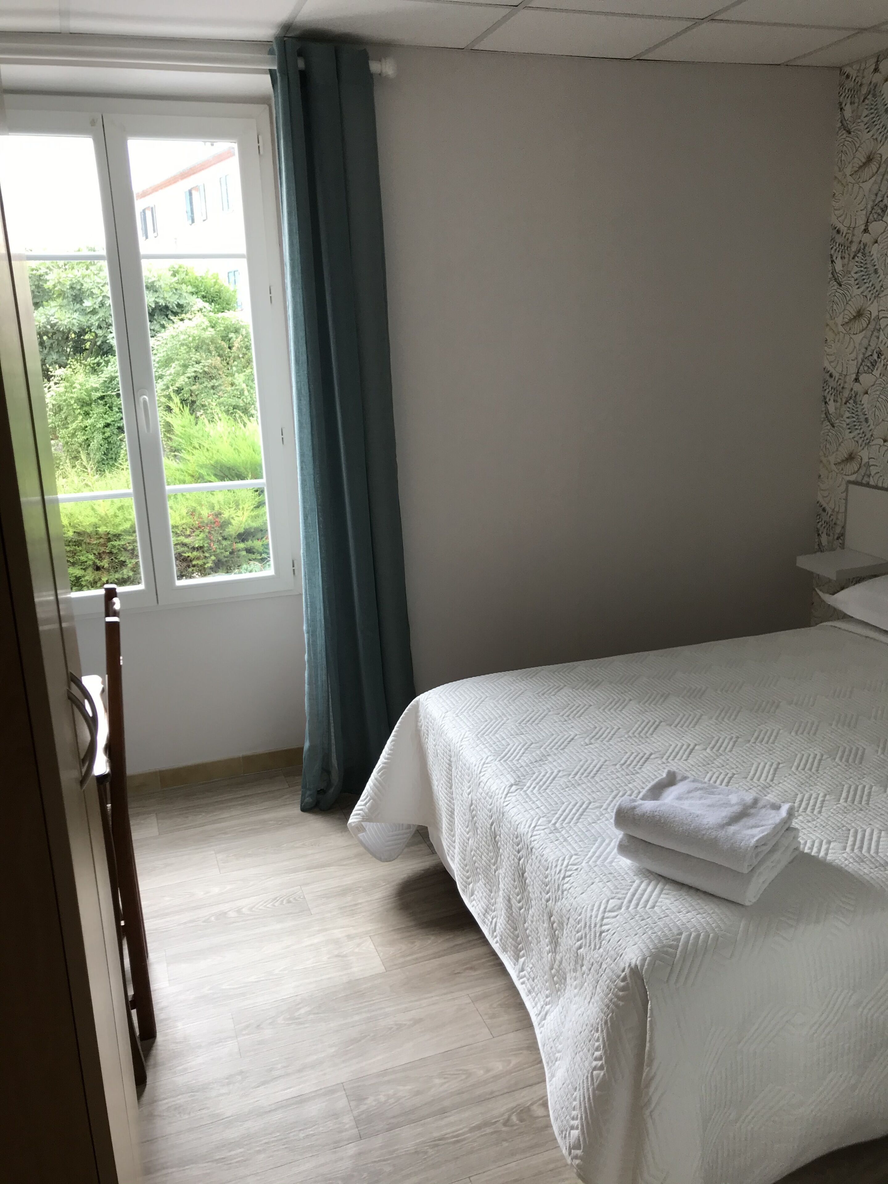 A hotel room with a bed, a window with curtains, and a view of greenery outside.