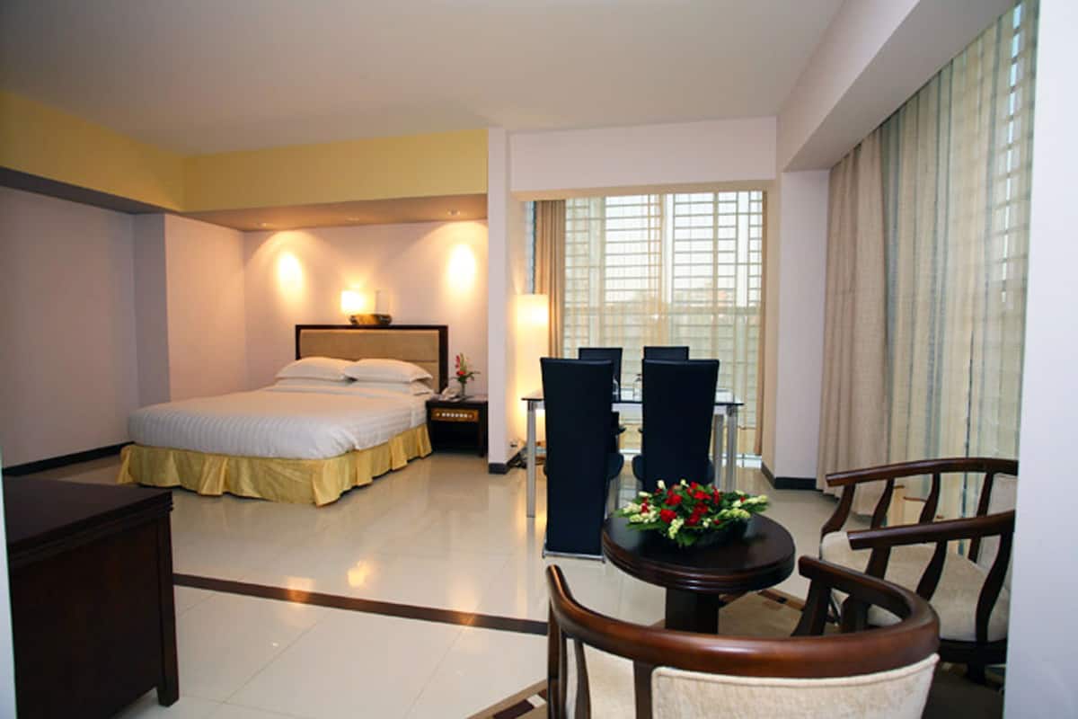 Habitación de hotel con cama, mesita de noche, lámpara, dos sillas, una pequeña mesa con flores y un amplio ventanal con cortinas.
