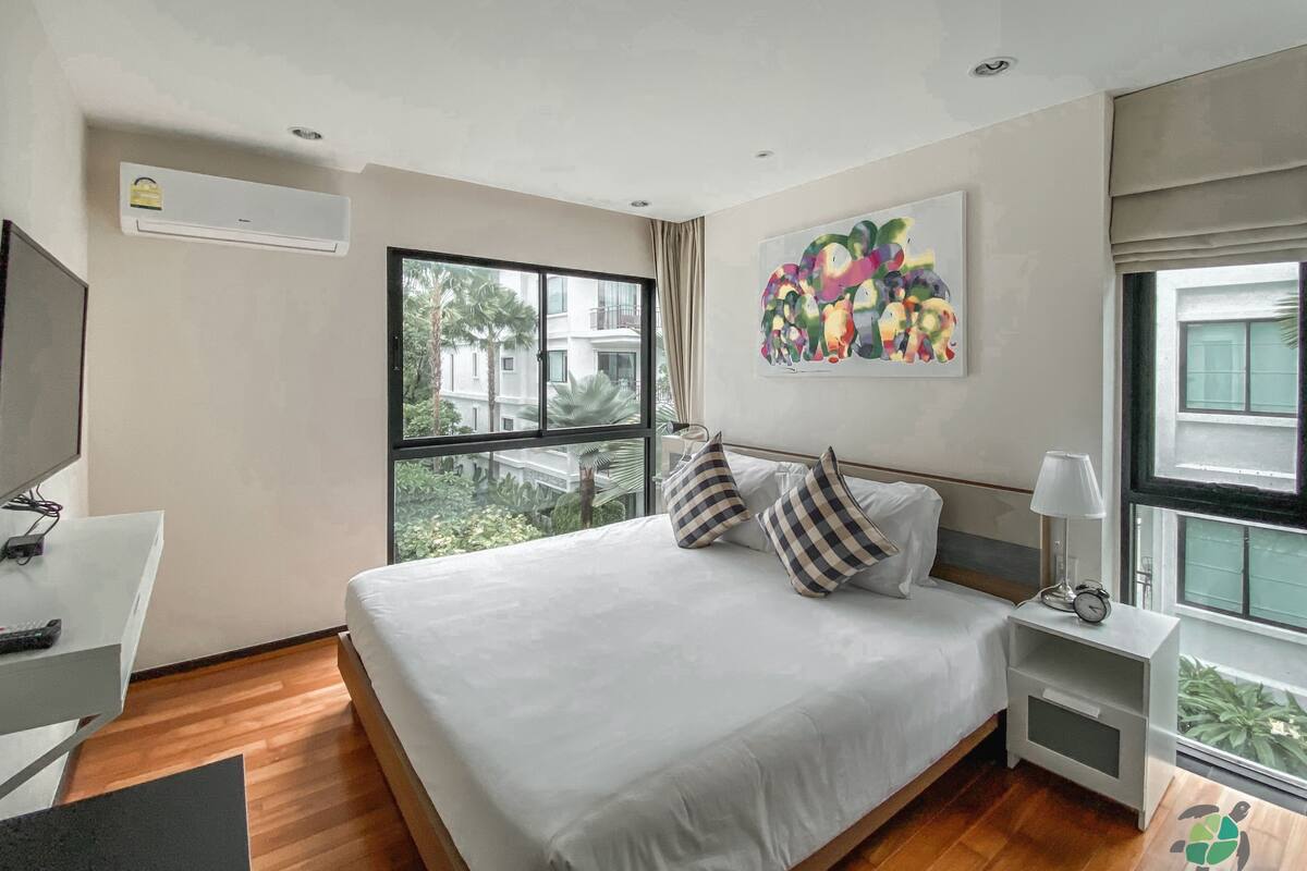Quarto de hotel com uma cama grande, uma escrivaninha, uma cadeira, uma televisão, uma janela com vista para edifícios e árvores, e um quadro na parede.