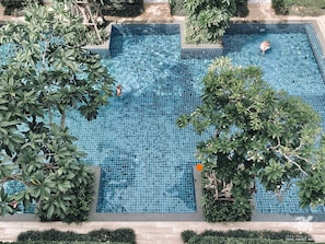 An aerial view of a rectangular swimming pool with blue mosaic tiles, surrounded by lush greenery and a tiled deck.