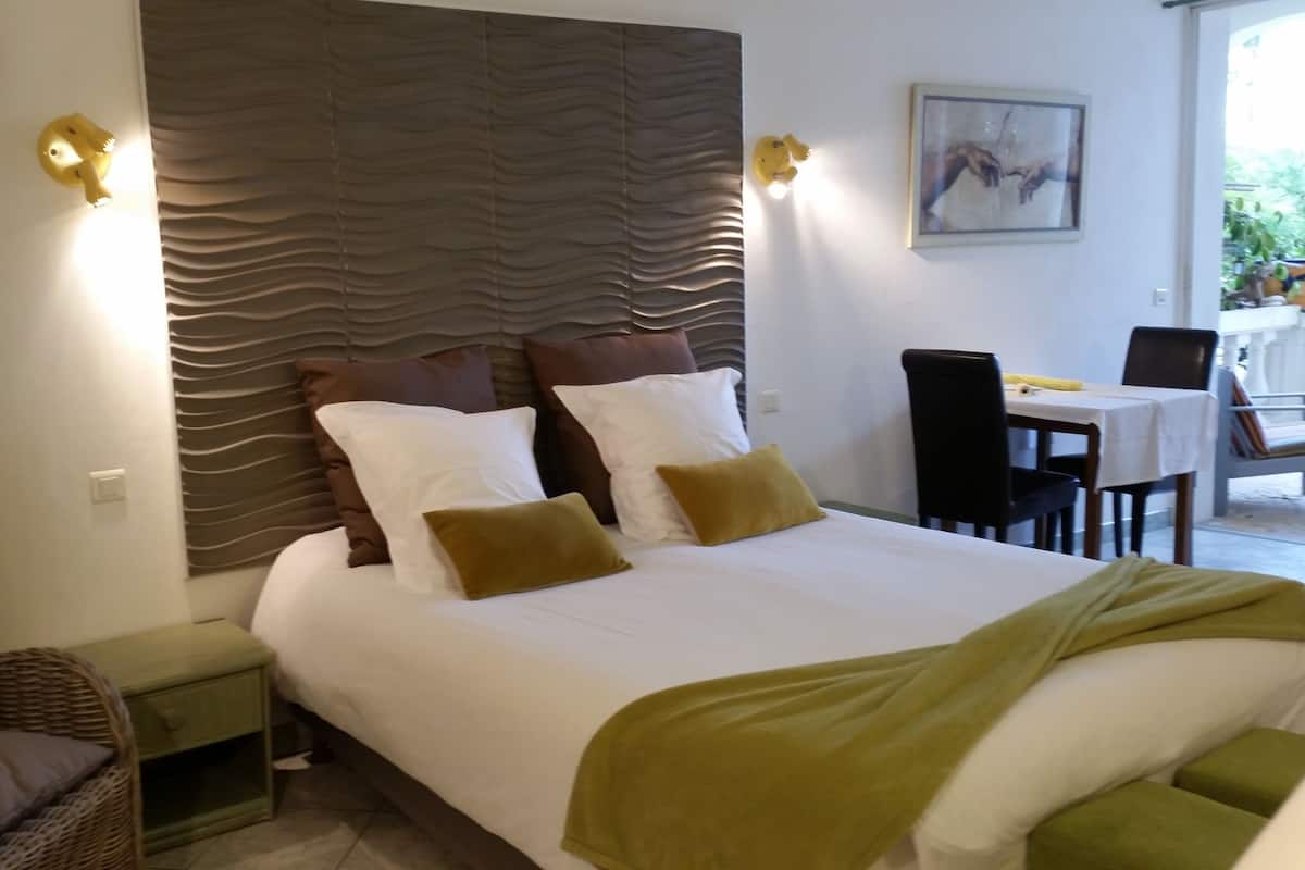 Une chambre d’hôtel avec un grand lit, une tête de lit au motif ondulé, une table de chevet verte, un fauteuil en osier et un coin repas avec une table et des chaises.