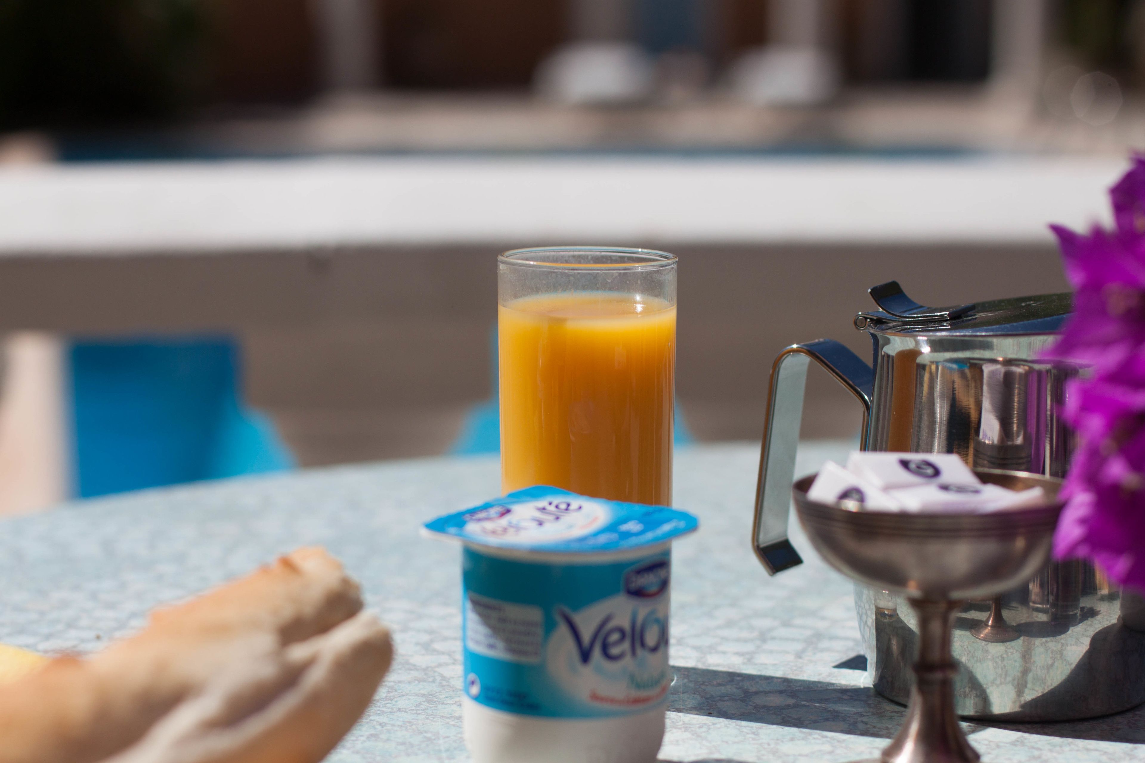 Une table avec un verre de jus d’orange, un pichet en métal, un bol rempli de serviettes en papier et un pot de yaourt.