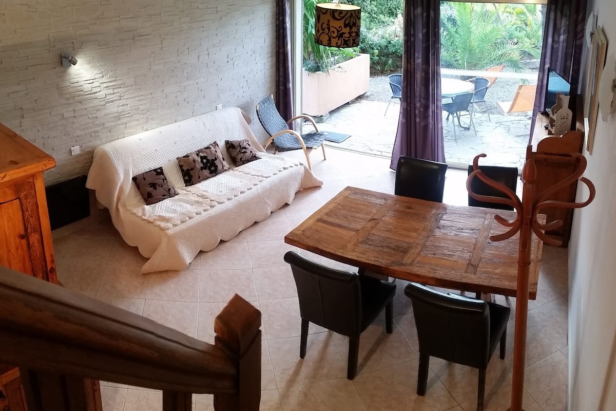 Un salon chaleureux avec un canapé blanc, une table à manger en bois et des chaises. Un escalier mène à l’étage supérieur.