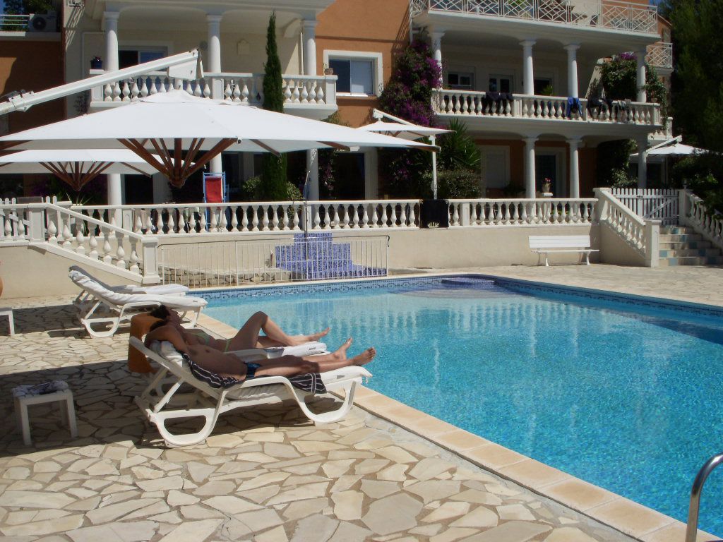 Zona de piscina con sillones y sombrillas, una escalera que lleva a un balcón, y un edificio con fachada blanca y balcones.