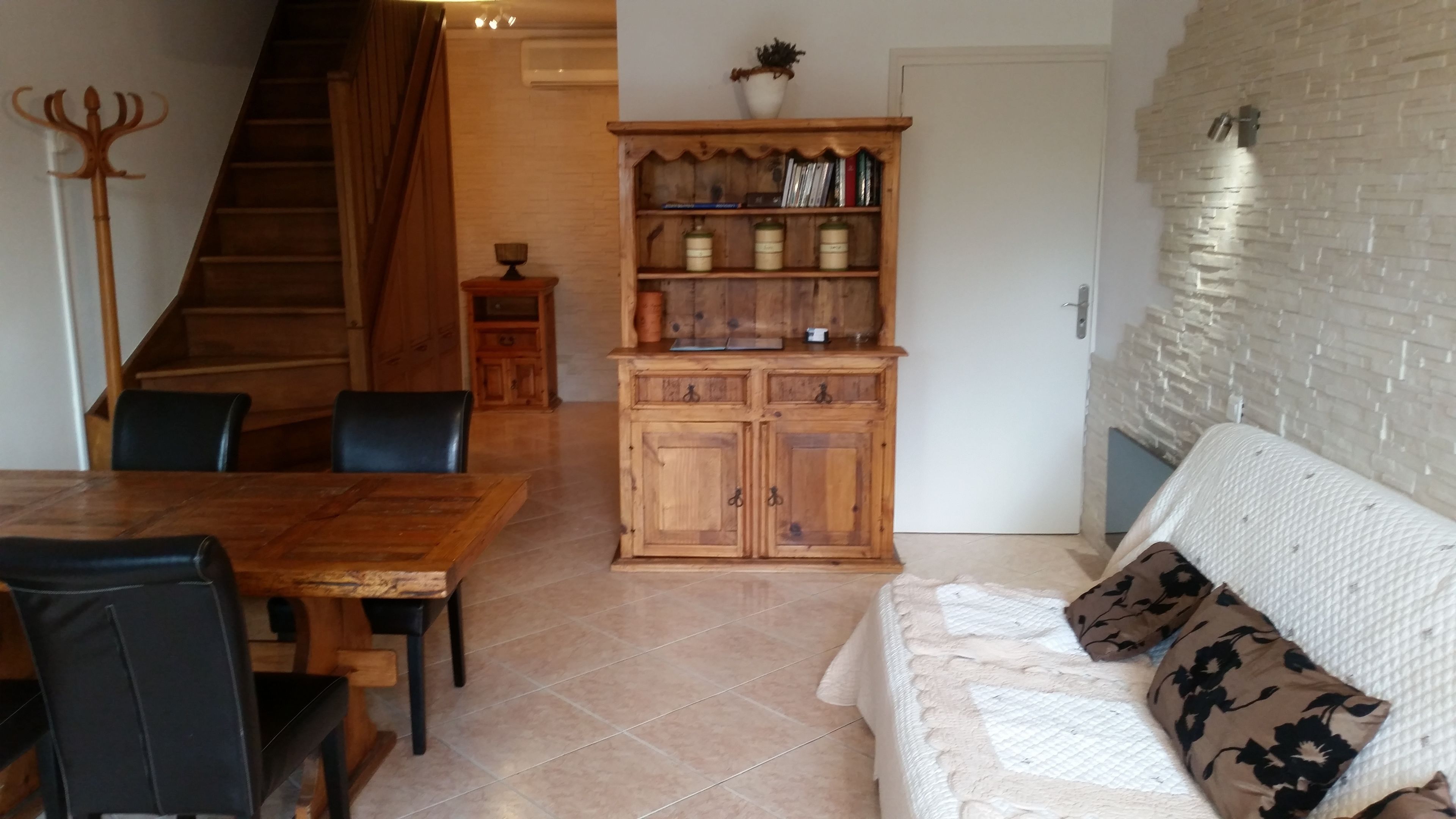 A dining area with a wooden table and chairs, a wooden cabinet with shelves, a staircase, and a white textured sofa with patterned cushions.