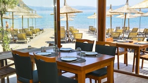A beachside restaurant with wooden tables and chairs, thatched umbrellas, and a view of the sea.