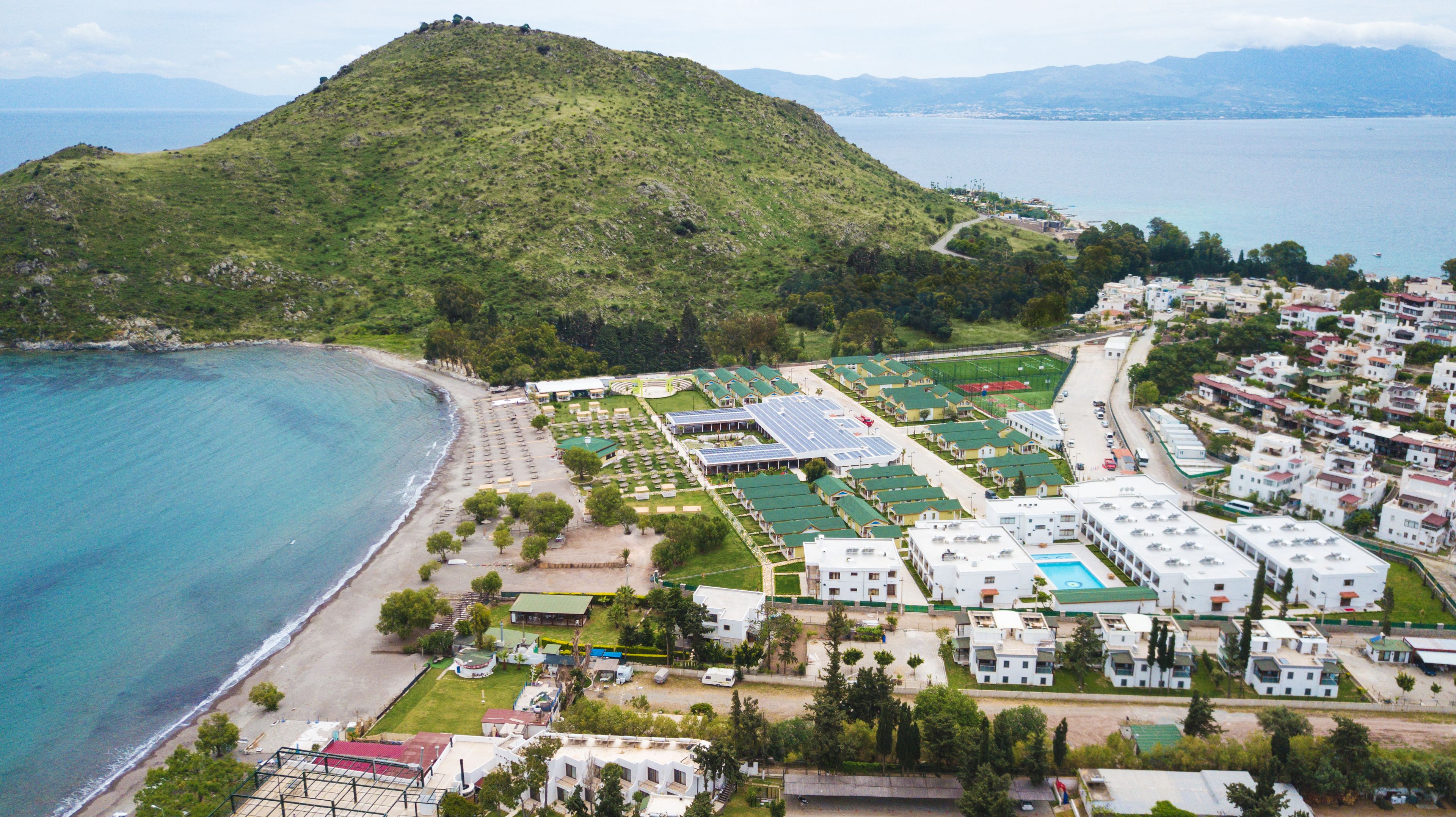 An aerial view of a coastal resort with a beach, a hill, and a cluster of modern buildings with solar panels.