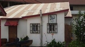 Ein kleines Haus mit Wellblechdach, zwei vergitterten Fenstern und einem kleinen Garten mit Pflanzen und einer Statue.