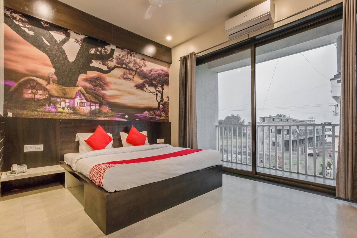 A modern hotel room with a large bed, a painting of a landscape, a balcony with a view of buildings, and an air conditioning unit.
