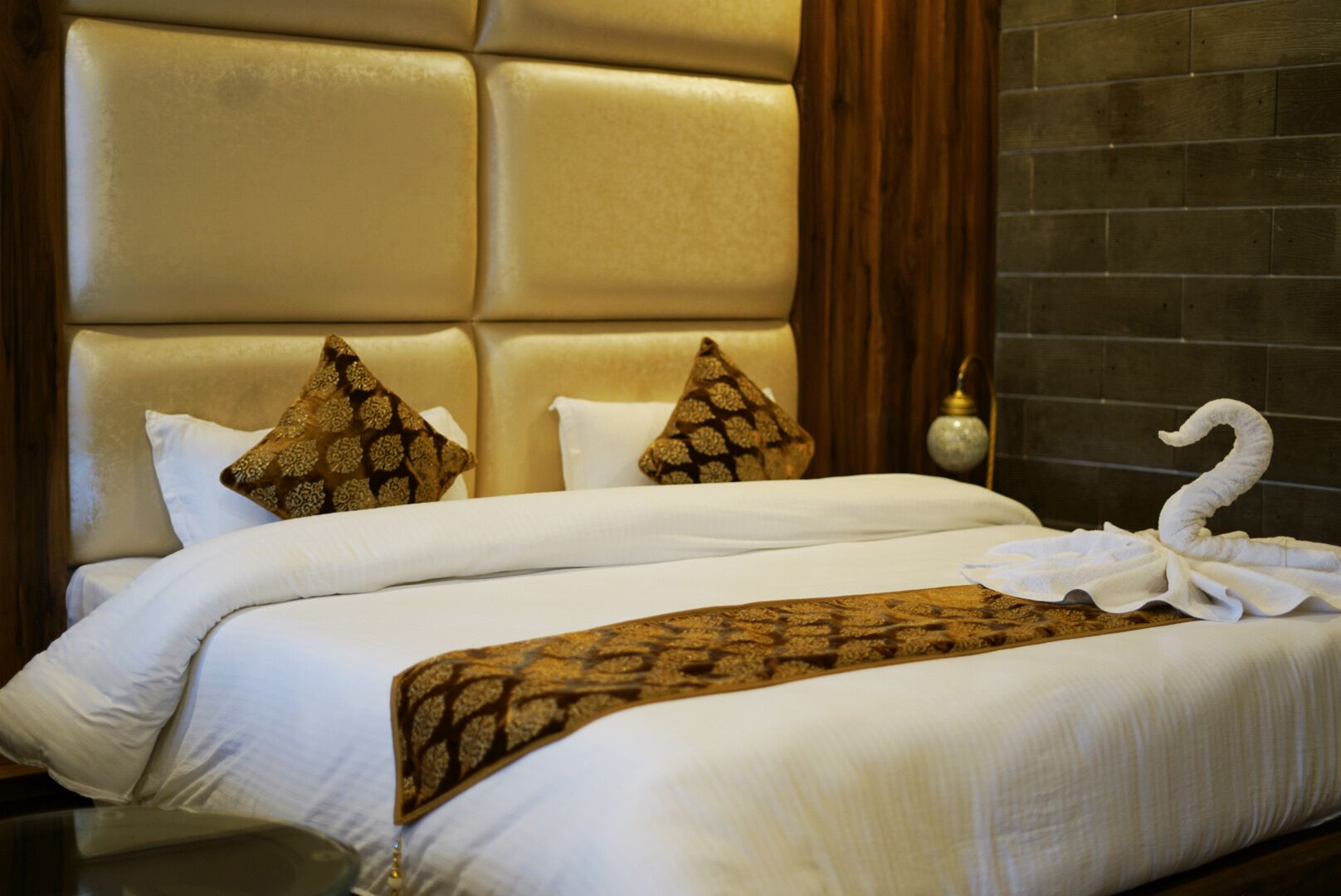 A hotel room with a large padded headboard, two white beds with matching pillows, and a decorative towel on the bed.