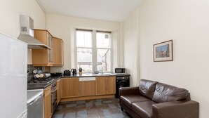 A compact kitchen with a stove, microwave, and dishwasher. There's a window with a view of buildings outside. A brown leather sofa is against the wall. - Silver Lining Lochrin Terrace Apartment (Edinburgh)