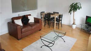 Ein Wohnzimmer mit braunem Ledersofa, einem Glastisch, einem Esstisch mit Stühlen, einer Topfpflanze und einem Fernseher.
