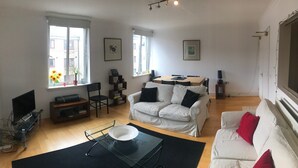 Ein Wohnzimmer mit einem weißen Sofa, einem Couchtisch, einem Fernseher und einem Esstisch mit Stühlen.