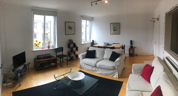 Ein Wohnzimmer mit einem weißen Sofa, einem Couchtisch, einem Fernseher und einem Esstisch mit Stühlen.