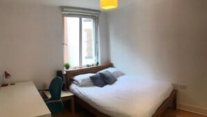 Ein Schlafzimmer mit einem Bett, einem Schreibtisch, einem Sessel, einem Fenster und einer kleinen Pflanze.