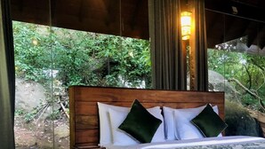 A wooden-framed bed with white bedding and green pillows, a small wooden nightstand, and a view of a natural outdoor area through a glass wall. - Arangala Forest Lodge (Naula)