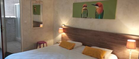 Un cuarto de hotel con una cama grande, dos lámparas de noche, un cabecero de madera, un taburete pequeño y un cuadro de pájaros en la pared.