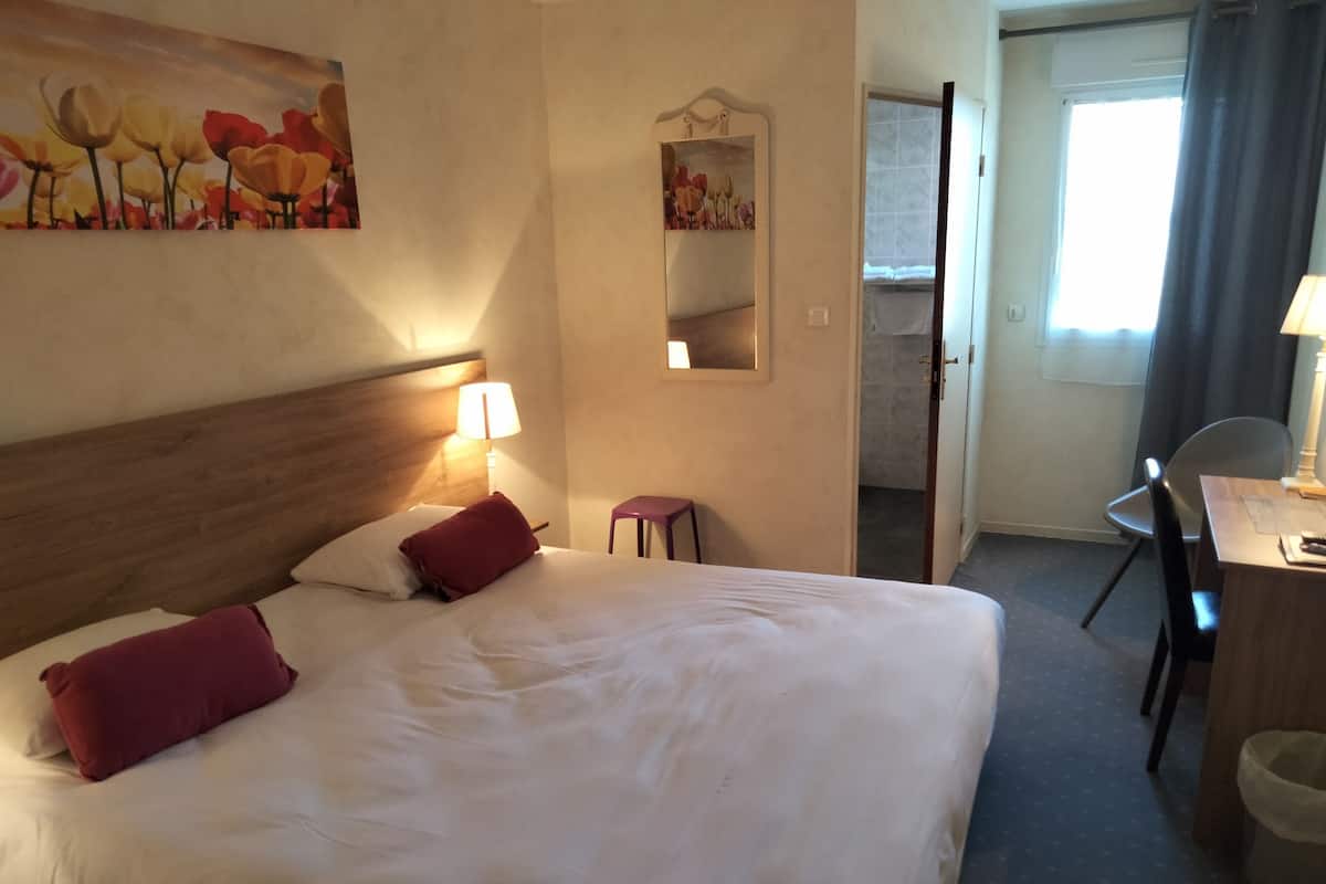 Een hotelkamer met een groot bed, een bureau met stoel, een klein krukje, een spiegel en een schilderij aan de muur.