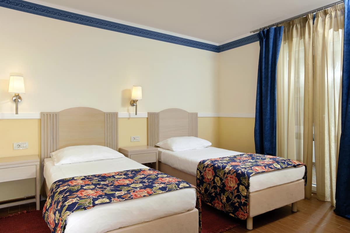 Quarto de hotel com duas camas de solteiro, cada uma com colcha florida, luminárias laterais e uma janela com cortinas azuis.