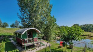 A green wooden cabin with a porch, a swing, and a pond in the background. - L'ecrin du Segala - La roulotte (Tauriac-de-Naucelle)