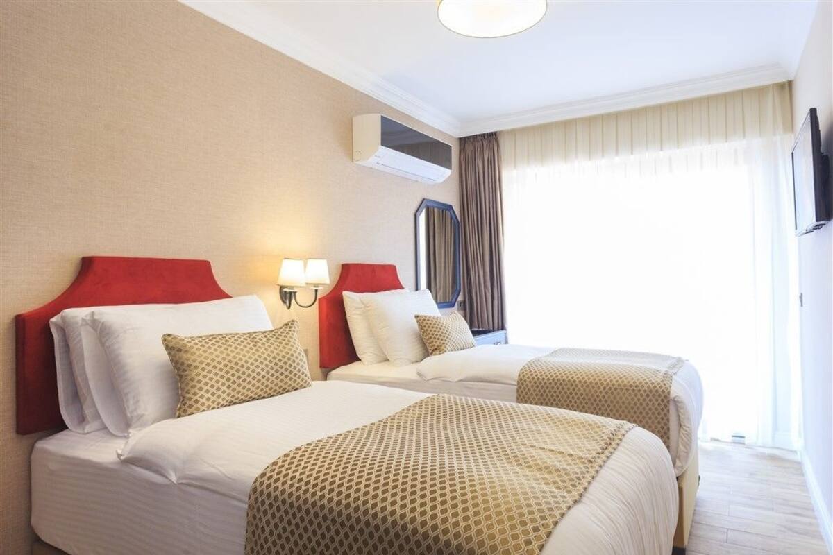 Une chambre d’hôtel avec deux lits, une tête de lit rouge, un climatiseur fixé au mur et une grande fenêtre avec des rideaux.