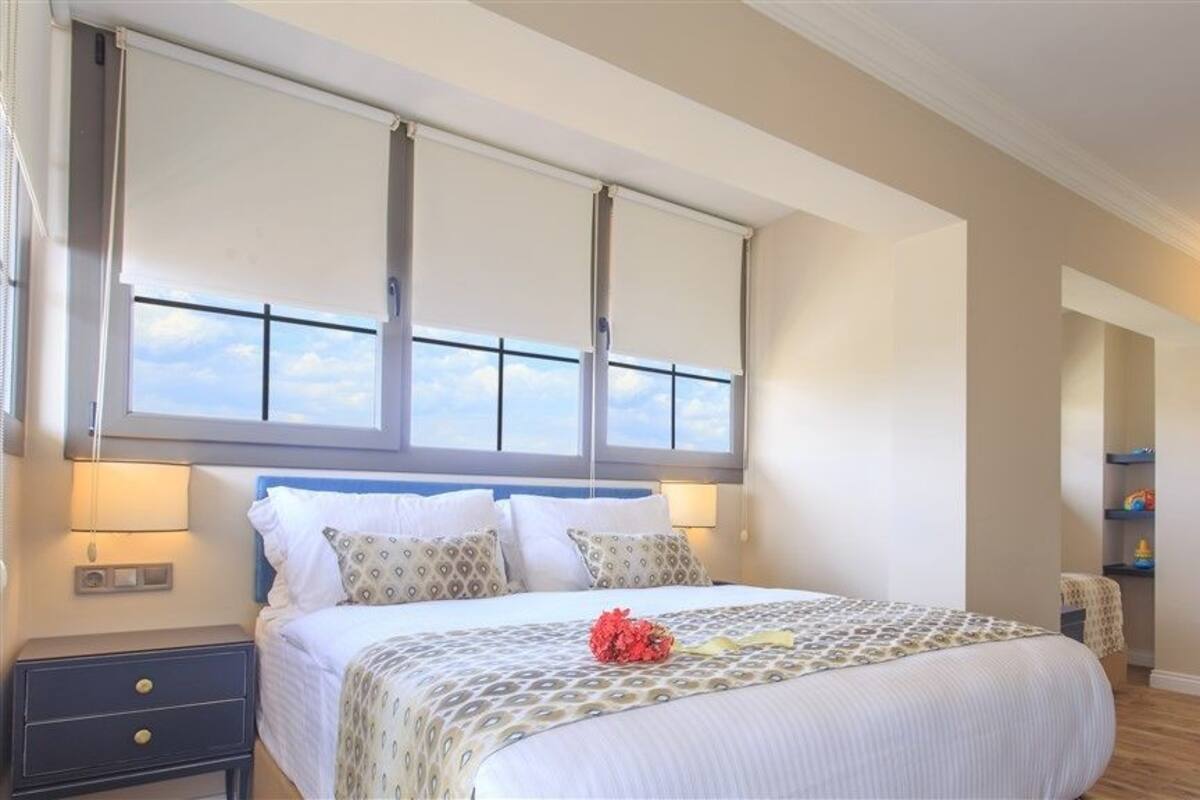 Une chambre à coucher avec un grand lit, une fenêtre avec des stores, une table de chevet et une commode.