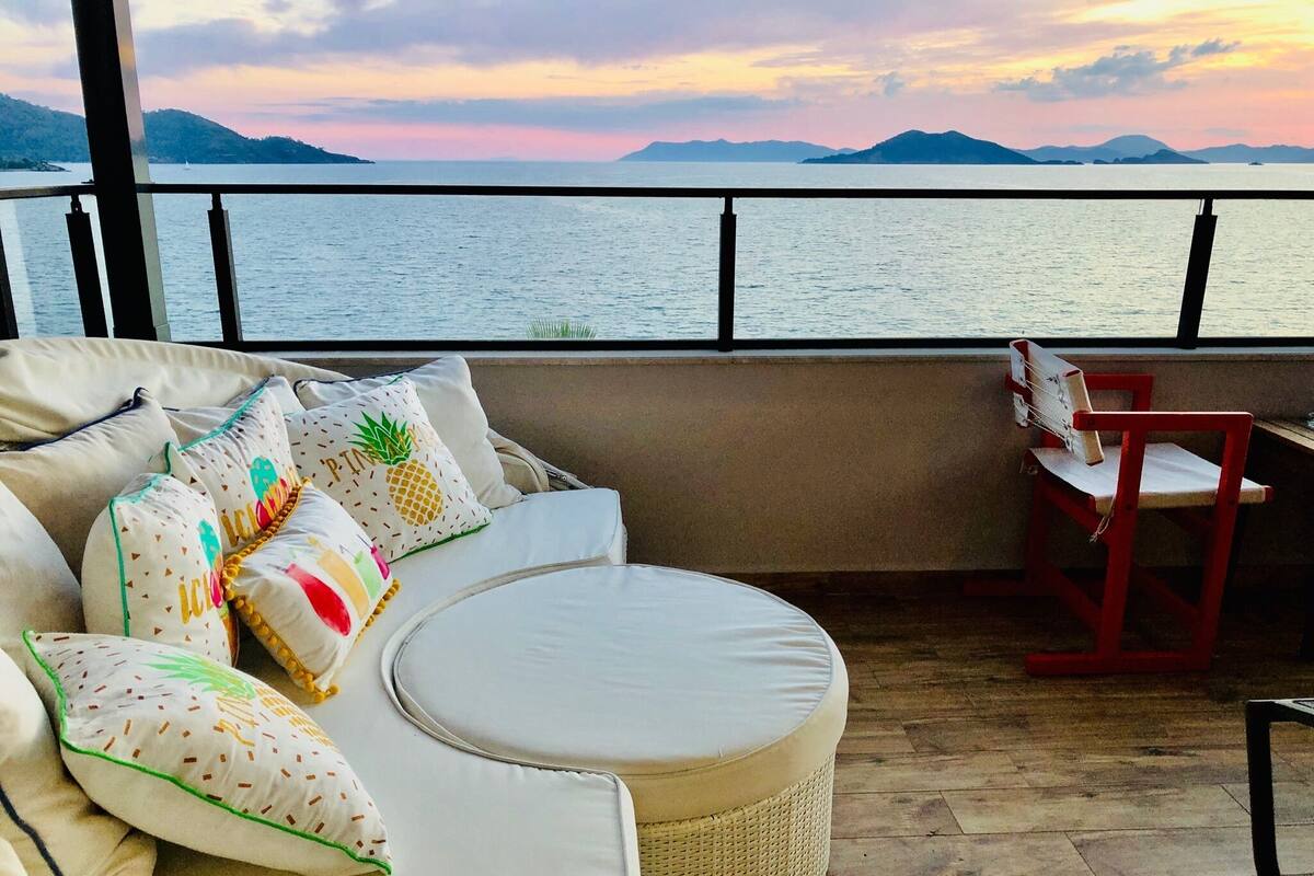 Un balcon avec un canapé et un pouf, une chaise rouge, et une vue sur la mer et les montagnes.