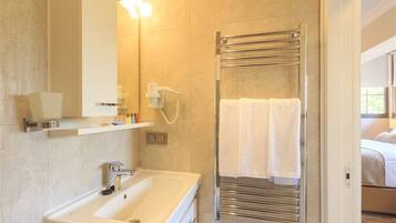 Un baño moderno con un lavamanos blanco, un perchero con dos toallas, un cabezal de ducha manual y un espejo con luces.